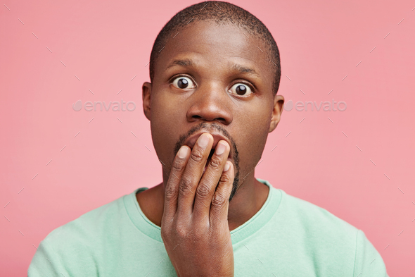 Frustrated puzzled shocked male with dark skin keeps hand on mouth, being surprised to make big mist