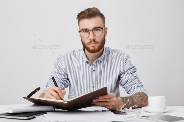 Portrait of confident male manager with tattooes, writes in diary plan for next week, drinks coffee,