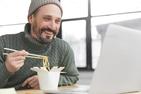 Positive middle aged bearded male eats pasta with chopsticks, has dinner break, watches interesting