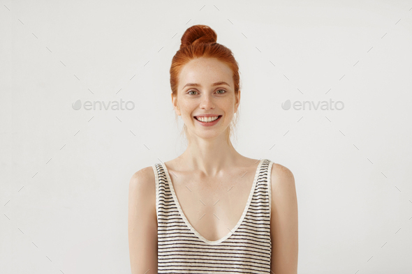 Emotional young female with red hair bun, wearing loose shirt, smiling cheerfully at camera after re