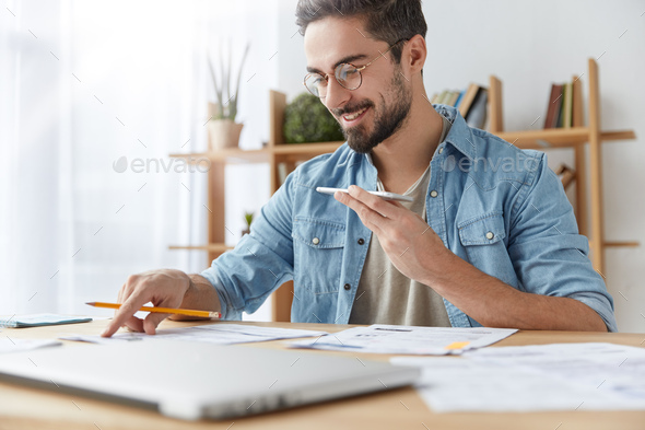Portrait of cheerful bearded man has voice call, speaks with friend and checks documents, has happy