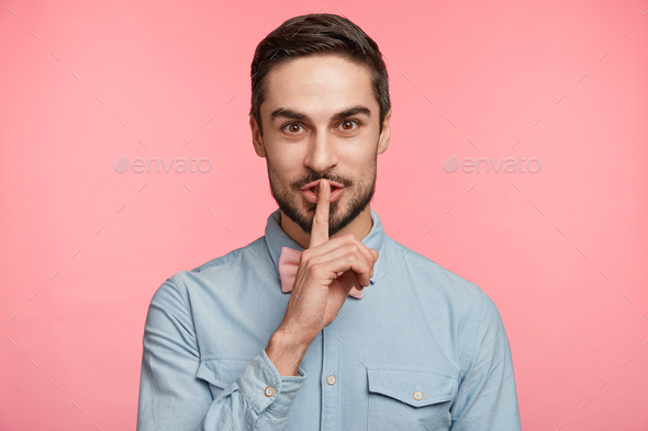 Attractive pleasant looking unshaven man keeps finger on lips, asks friends not to tell secret and w