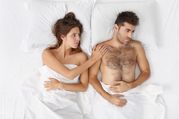 Attractive female cheering up her anxious man in bed. Bearded Caucasian male feeling frustrated becu - Stock Photo - Images