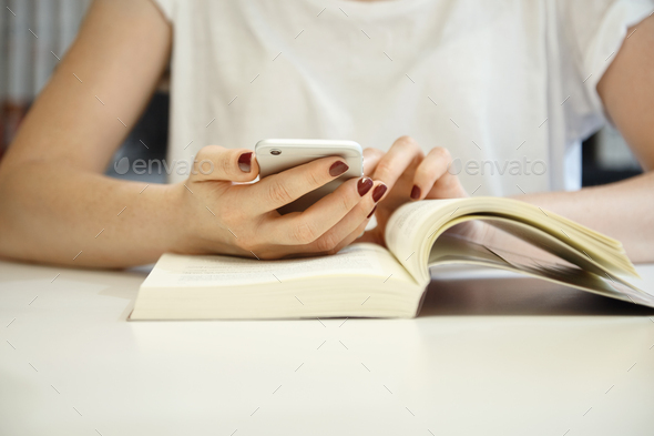 Cropped shot of female with neat manicure wearing white blouse with her hands on an open book, brows