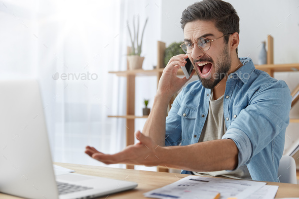 Annoyed male enterpreneur looks angrily into laptop computer, has conversation with business partner