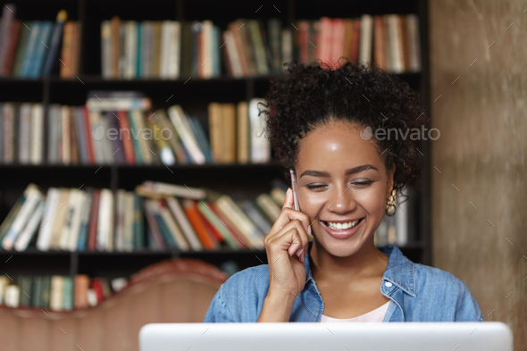 Beautiful student girl with charming smile talking on mobile phone while studying at college library - Stock Photo - Images