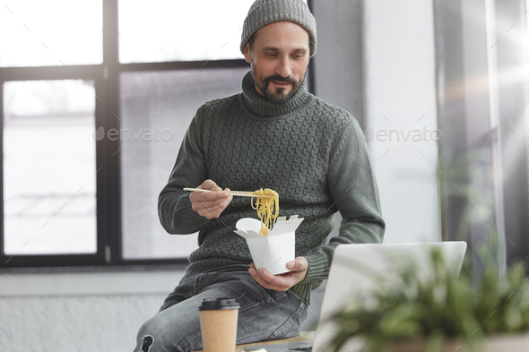 Delightful middle aged male with thick beard and mustache has dinner break in office, eats noddle wi