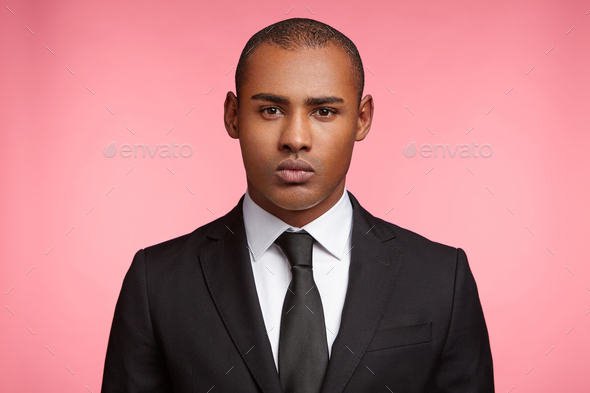 Headshot of serious tanned businessman wears formal suit and tie, looks directly into camera, thinks