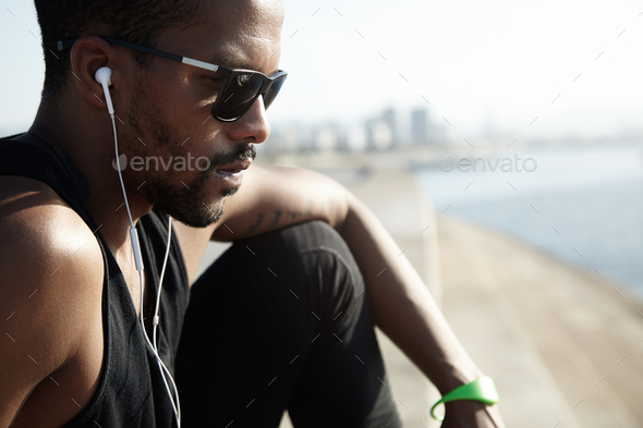 Cropped shot of pensive young handsome jogger in sunglasses sitting on top of stone stairs, dressed