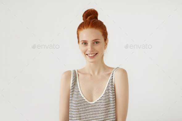 Positive young girl with ginger hair knot, wearing loose sailor shirt, having charming smile, isolat