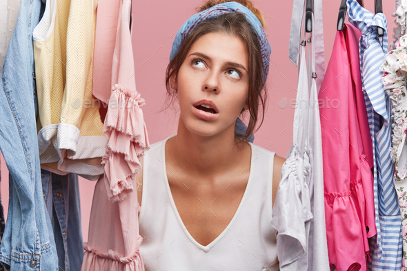 Portrait of good looking female with tired expression, looking through rack of clothes, doing shoppi