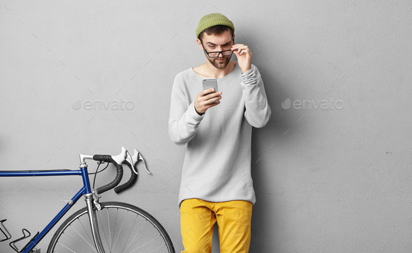 Picture of young bearded male wearing trendy hat and clothes holding eyeglasses while reading strang
