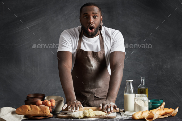 Shocked dark skinned male cook wears apron, kneads dough, being surprised to find out about deadline