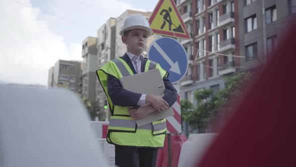 Portrait Thoughtful Little Boy Wearing Safety Equipment and Constructor Helmet Holding Building Plan alt