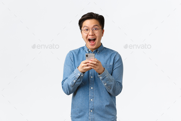 Excited happy asian man reacting cheerful at awesome news read online, holding mobile phone and