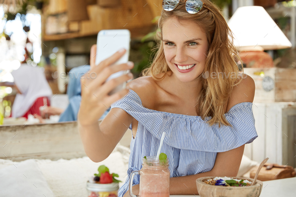 Glad young woman makes selfie on smart phone while has rest in cozy cafeteria, going to download pho