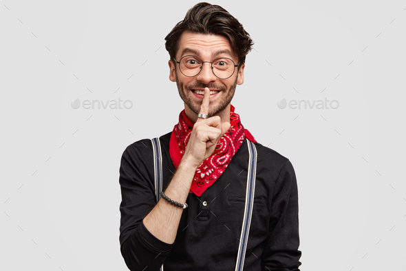 Happy delighted unshaven male in fashionable clothing, makes silence sign, asks to stop gossiping, i