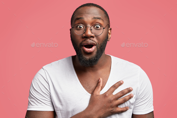 Studio shot of excited glad African American male model keeps hand on chest, looks with surprise and