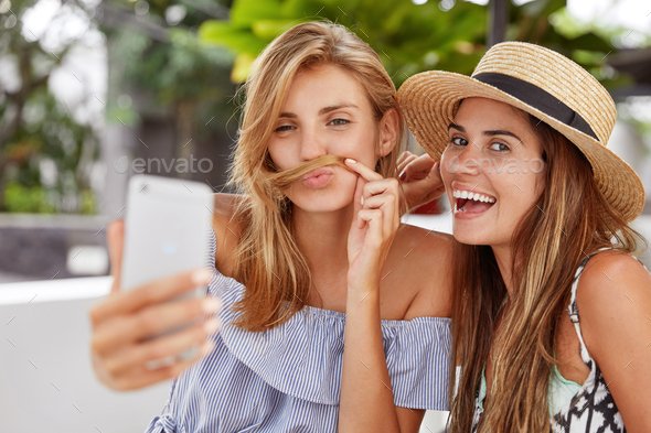 Joyful funny cute best female friends play fools at outdoor cafe, make grimace as pose for selfie in