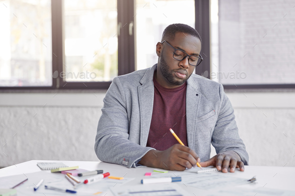 Serious dark skinned plump male entepreneur writes down ideas for creating productive strategy, sits