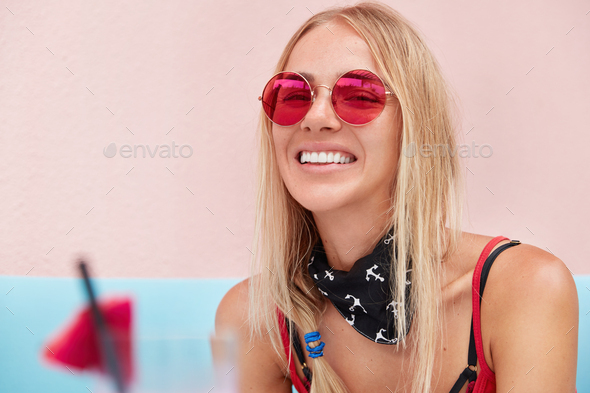 Horizontal shot of cute young woman wears fashionable red shades, has satisfied expression, poses ag