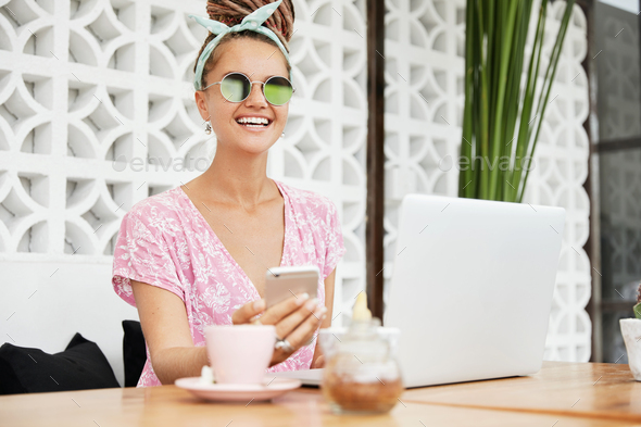 Stylish female blogger wears sunglasses, works remotely at laptop computer, develops personal websit
