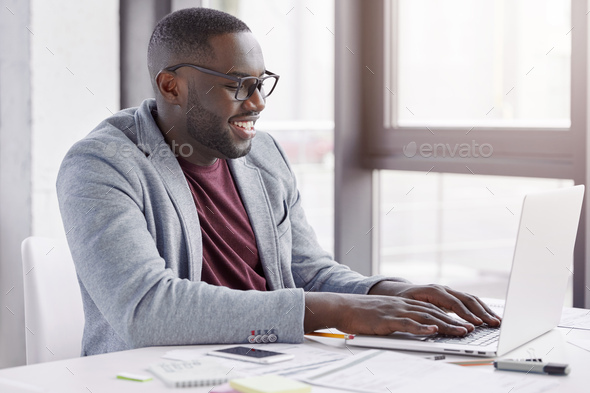 Pleased black Afro American male enterpreneur checks online database of finance report on laptop com