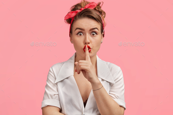 Studio shot of beautiful stunned housewife keeps index finger on lips, warns you to be silent as som