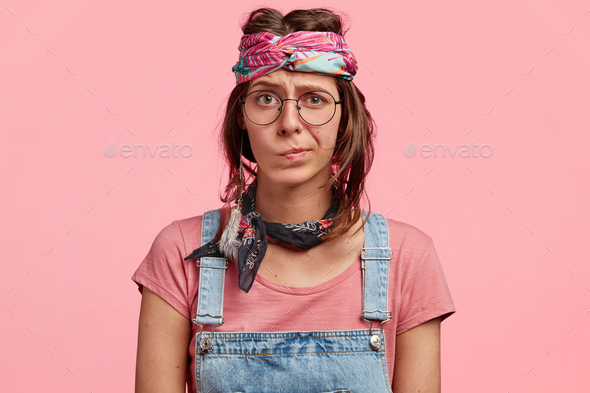 Attractive female hippie frowns face with doubt, has uncertain expression, wears stylish hippy`s clo