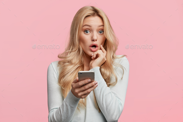 Beautiful female with blonde hair feels surprised as checks her email box on cell phone, wonders big