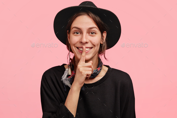 Adorable cheerful stylish hipster female shows shush sign, tells pleasant secret to best friend, ask