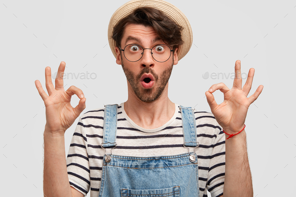 Horizontal portrait of surprised bearded handsome young hard working male farmer shows ok sign, reci