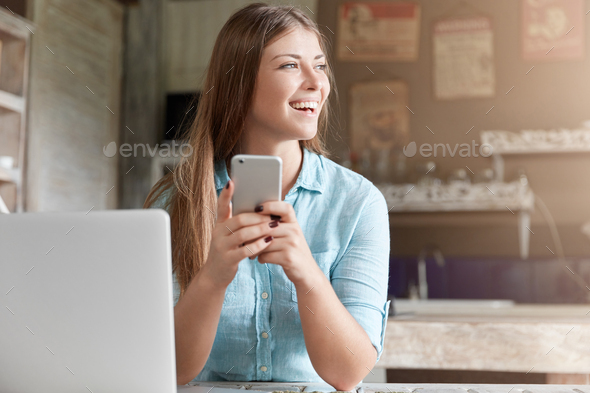 Happy beautiful woman with long straight hair reads text message om mobile work, sits in front of op