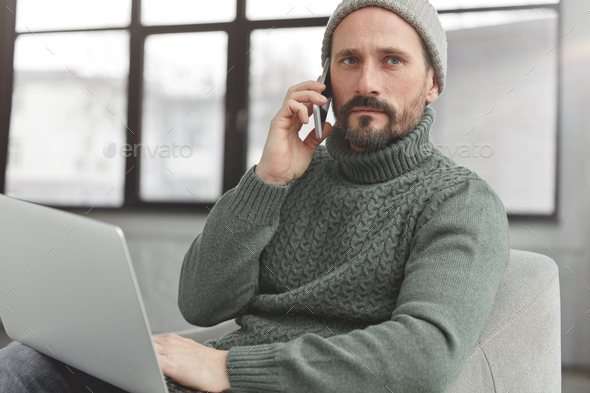 Bearded fashionable man with beard and mustache sits on comfortable sofa, calls someone via cell pho
