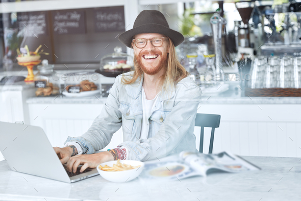 Smiling skilled male programmer has long hair, reddish beard and mustache, wears glasses, trendy hat - Stock Photo - Images