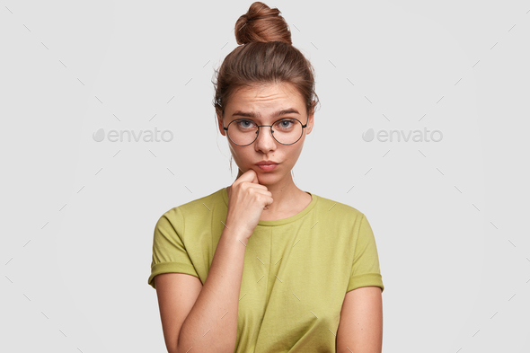 Horizontal portrait of lovely blue eyed female student with hair knot, wears big round spectacles, h
