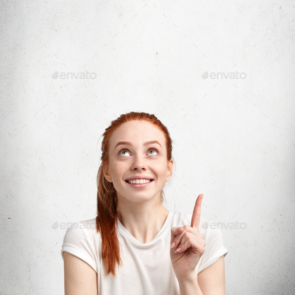 Positive red haired female with pleasant smile, dressed casually, points upwards at ceiling, adverti