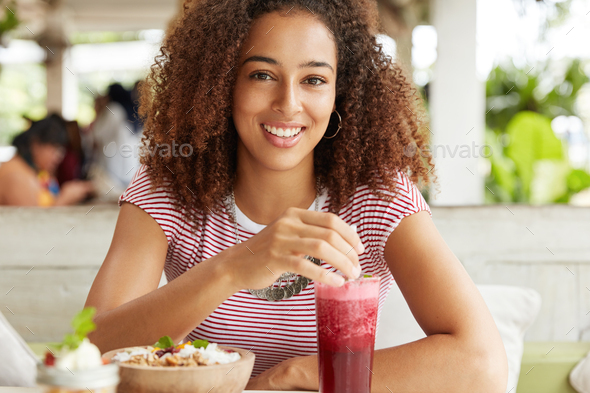 Beautiful African American female has curly hair, broad smile, sits at outdoor cafe, has dinner brea