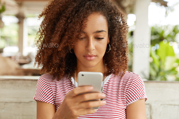 Serious dark skinned female reads income message with serious expression, checks email online on sma