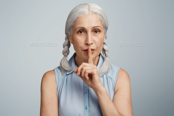 Keep secret, please. Elderly woman with two grey plaits holds finger on lips, shows silence sign, as