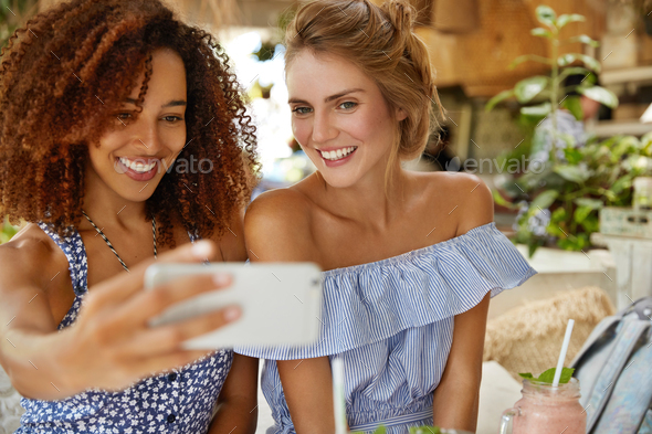Positive female best friends spend time together at cafe, pose at camera of modern cell phone, make