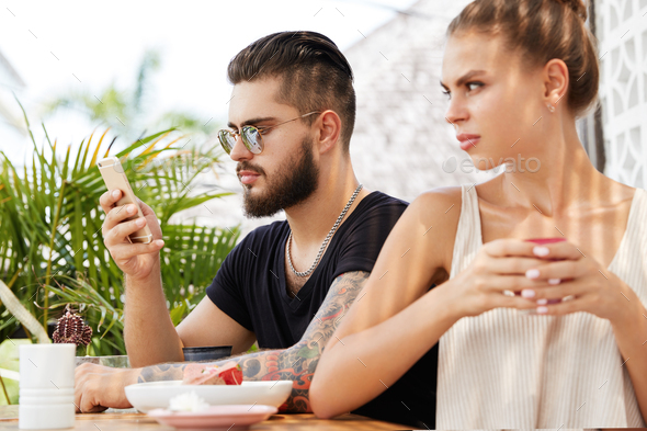 Displeased jealous female sits back to her boyfriend who messages on smart phone and doesn`t pay att
