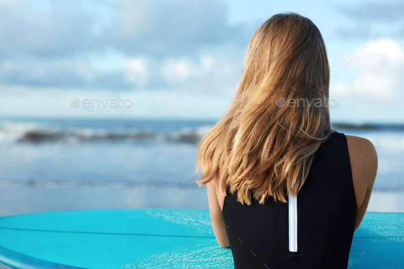 Unrecognizable female with straight blonde hair sits back at camera with surf board, spends spare ti