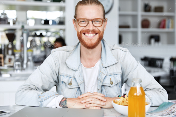 Pleased smiling hipster with ginger beard and mustache, wears spectacles, sits at table, has dinner