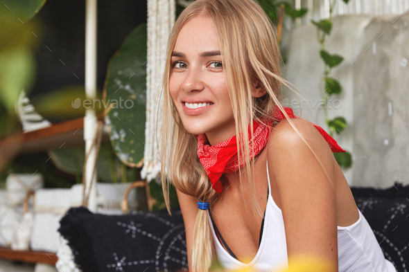 Fashionable delighted female wears tank top and bandana on neck, sits on comfortable couch at cozy c