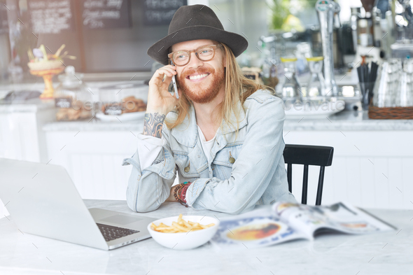 Cheerful hipster guy with long hair, dressed in fashionable denim jacket and black hat, communicates