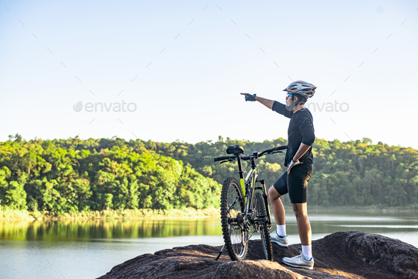Mountain cyclists stand on the top of the mountain with the bike and point the finger in front.