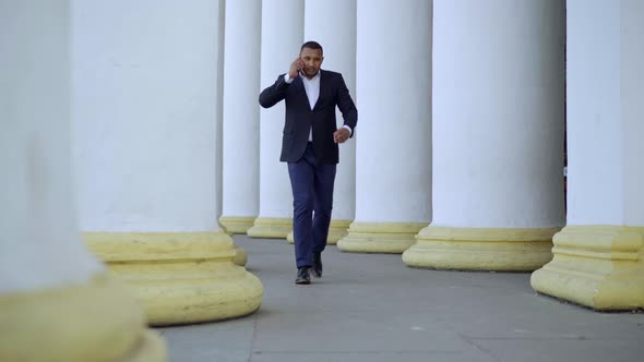 Wide Shot of Busy African American Businessman Talking on the Phone Walking Between White Columns alt