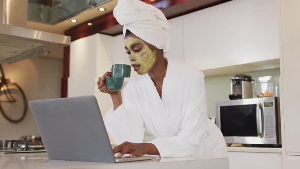 African american attractive woman with applied face mask drinking coffee and using laptop in kitchen alt