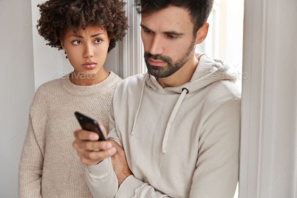 Attractive man wears hoodie, ignores live communication with wife, focused in modern smart phone, re